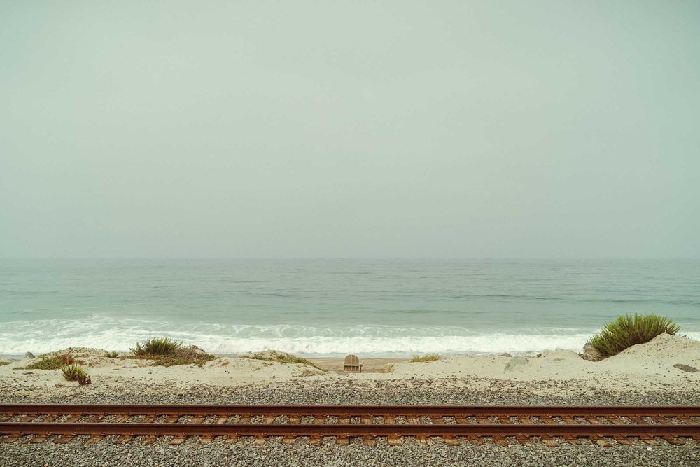 Minimalist coastal landscape with railroad tracks running parallel to a misty beach on an overcast day.