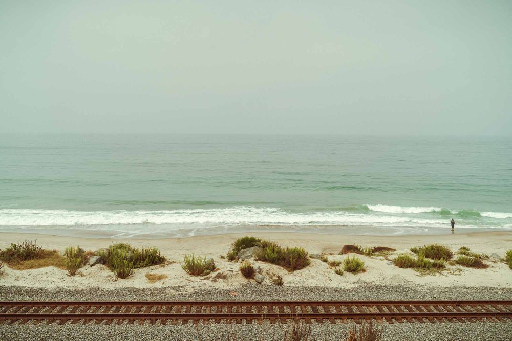Railroad tracks run parallel to a misty beach with sand dunes and gentle waves rolling onto the shore.