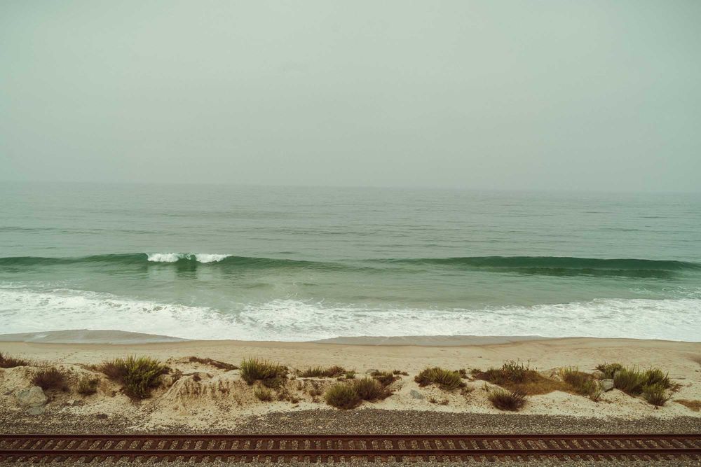 Gentle waves roll onto a sandy beach with dunes and railroad tracks under an overcast green-tinted sky.