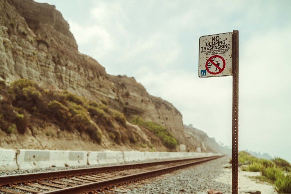 Railroad tracks run along steep coastal cliffs with a no trespassing sign visible in the foreground.