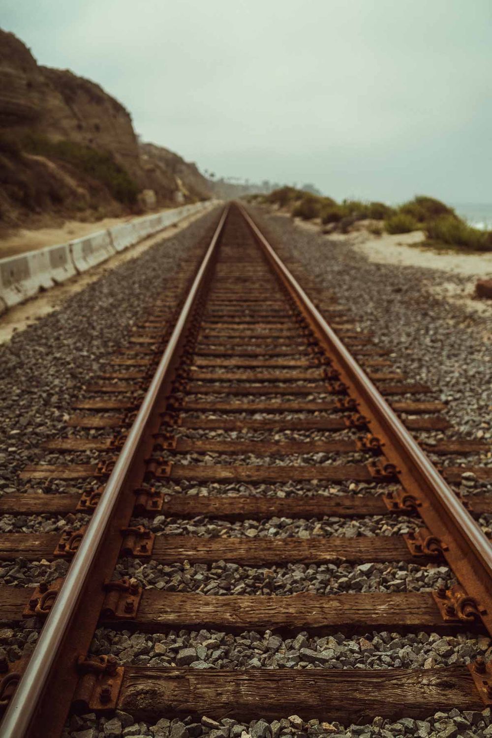 Railroad tracks stretch into the distance along a coastal cliff with wooden ties and gravel ballast under cloudy skies.
