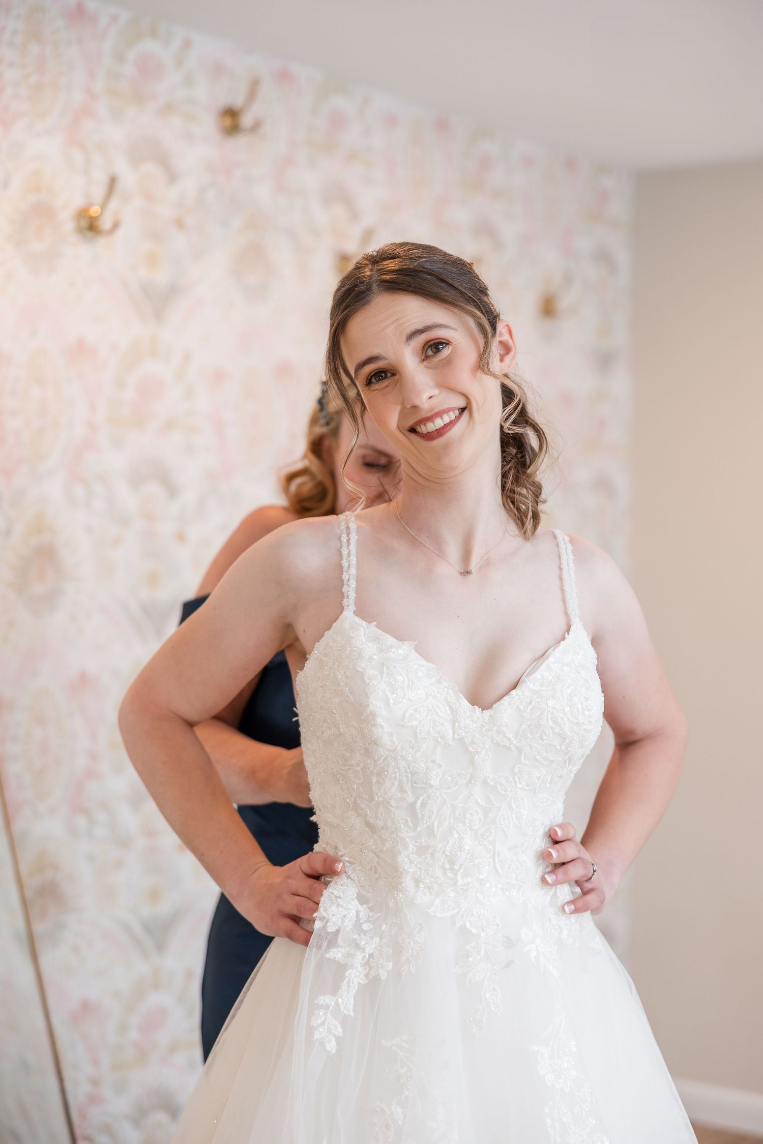 A bride in a white lace wedding dress gets ready in a room with floral wallpaper.