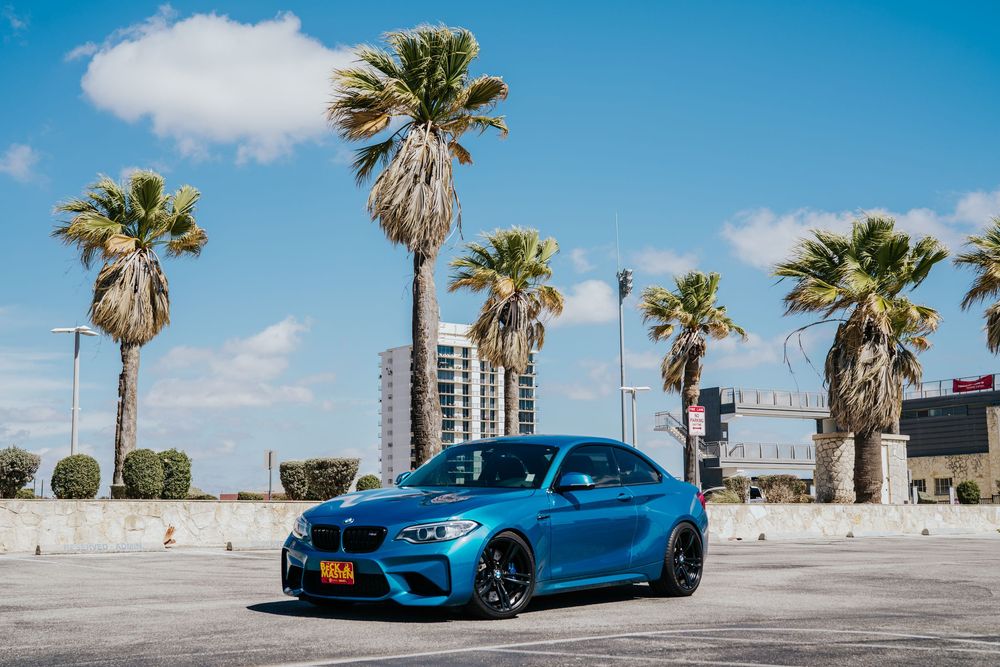 A bright blue BMW M2 sports car parked on a beachfront promenade with palm trees and clear skies in the background.