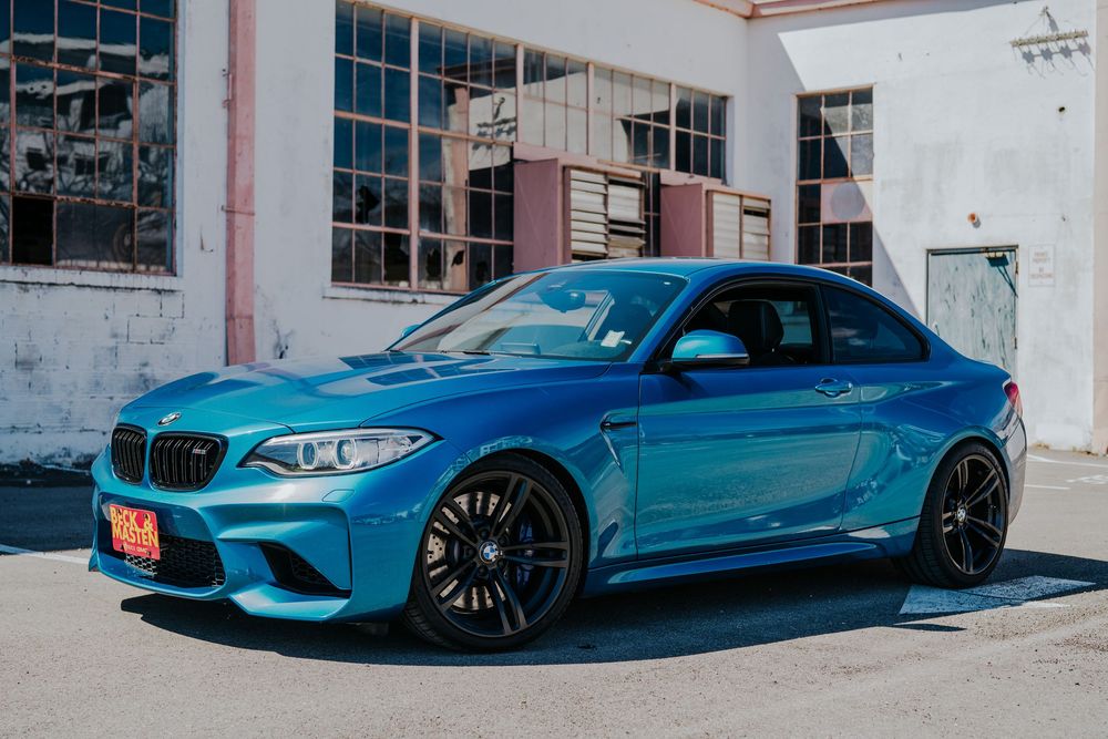 A metallic blue BMW M2 sports car with custom wheels parked in front of an industrial building.