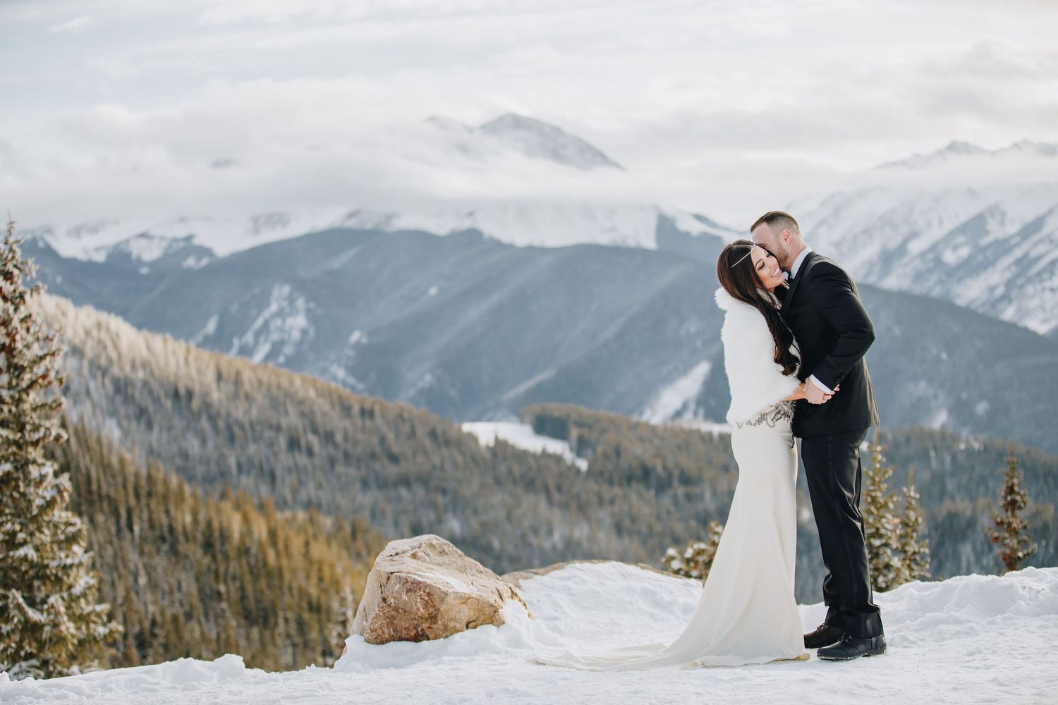 Couple shares an intimate moment on a snowy mountain peak with dramatic mountain ranges and evergreens in the background.