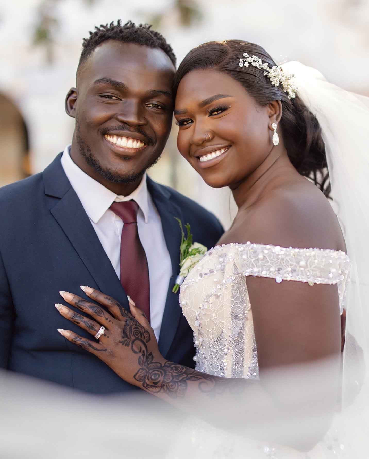 Smiling newlywed couple poses outdoors, bride wearing off-shoulder lace gown with veil.