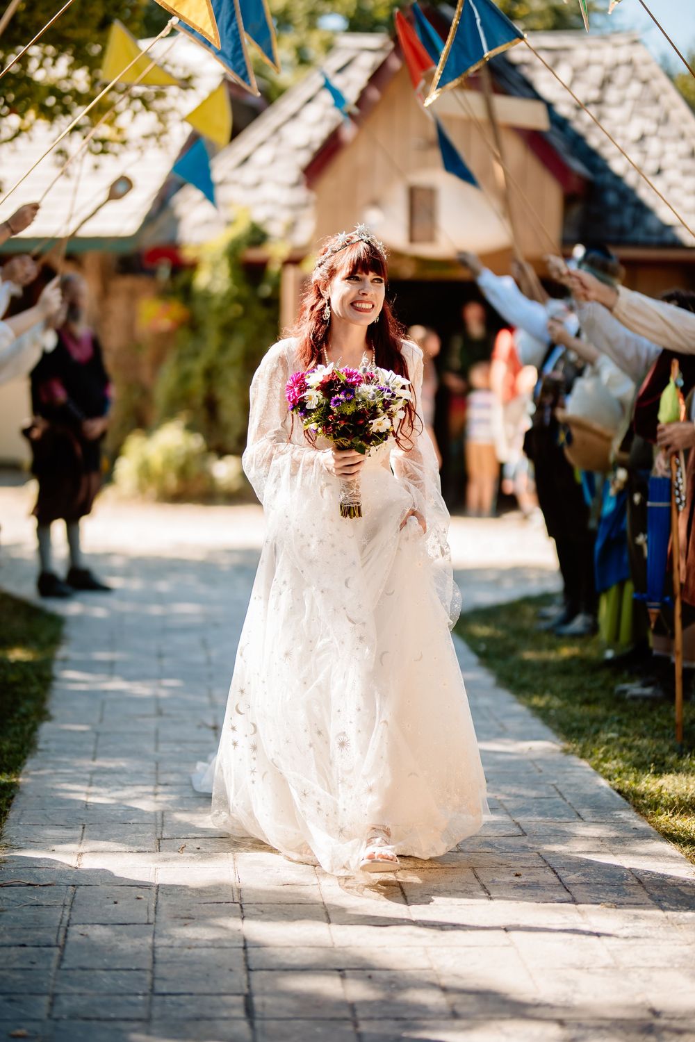 Radiant bride walks down a sunlit outdoor aisle holding a colorful bouquet at a Renaissance-themed wedding.