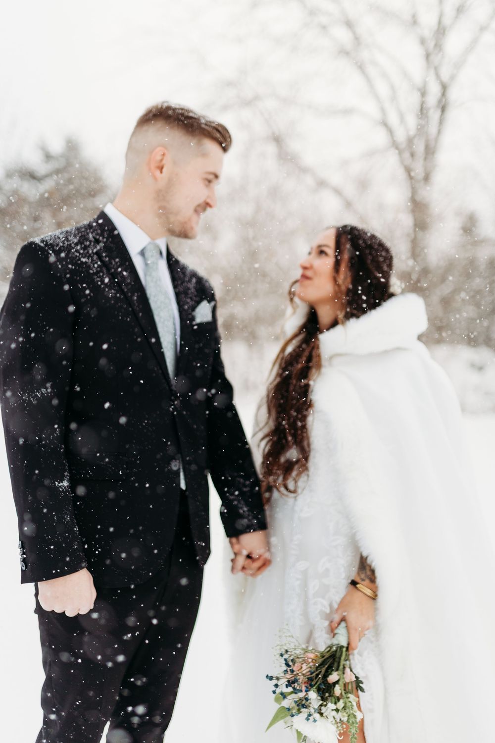 Bride in white fur cape and groom in black suit hold hands smiling in snowy winter wedding.