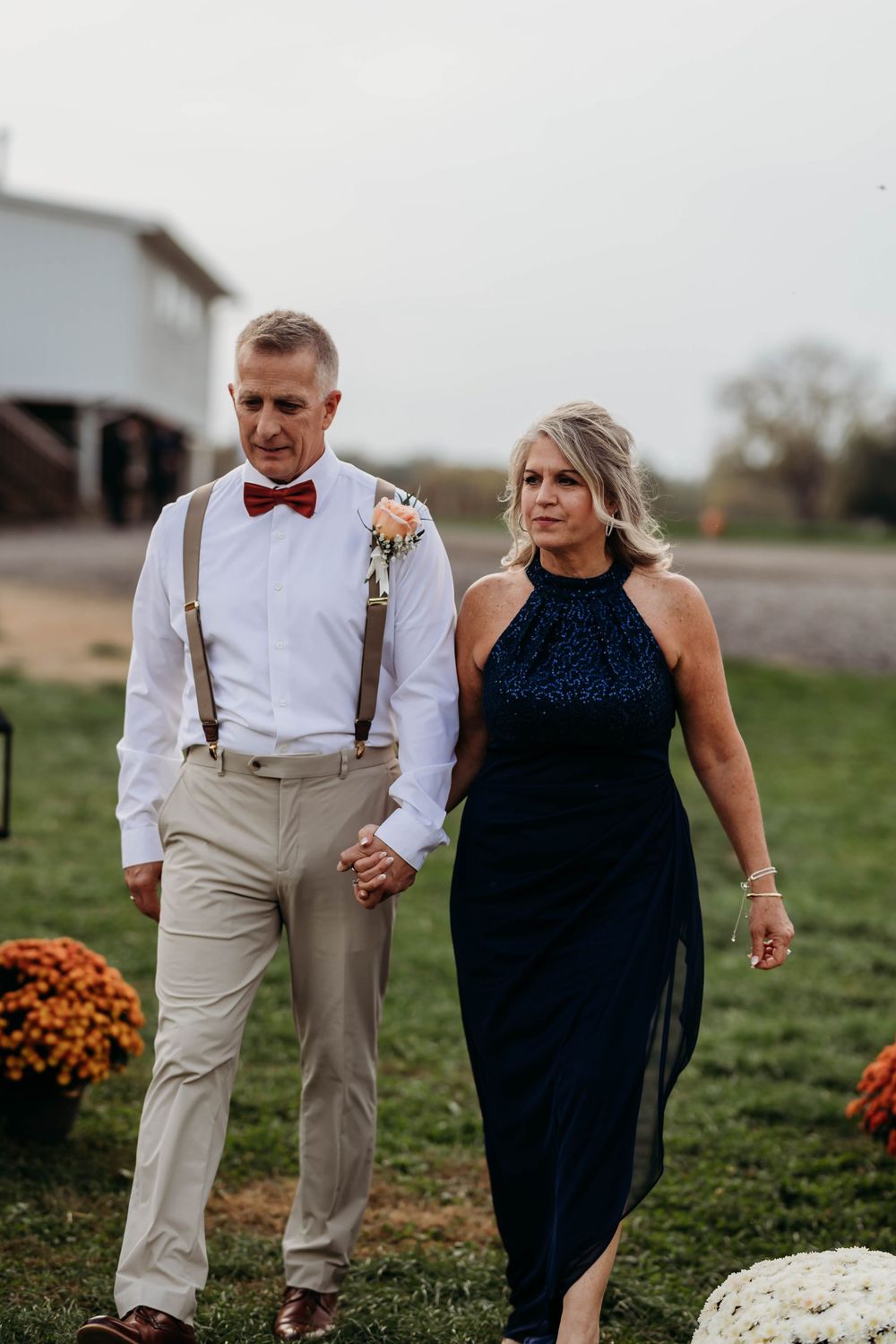 Groomsman in suspenders and red bow tie escorts woman in navy gown down the aisle.