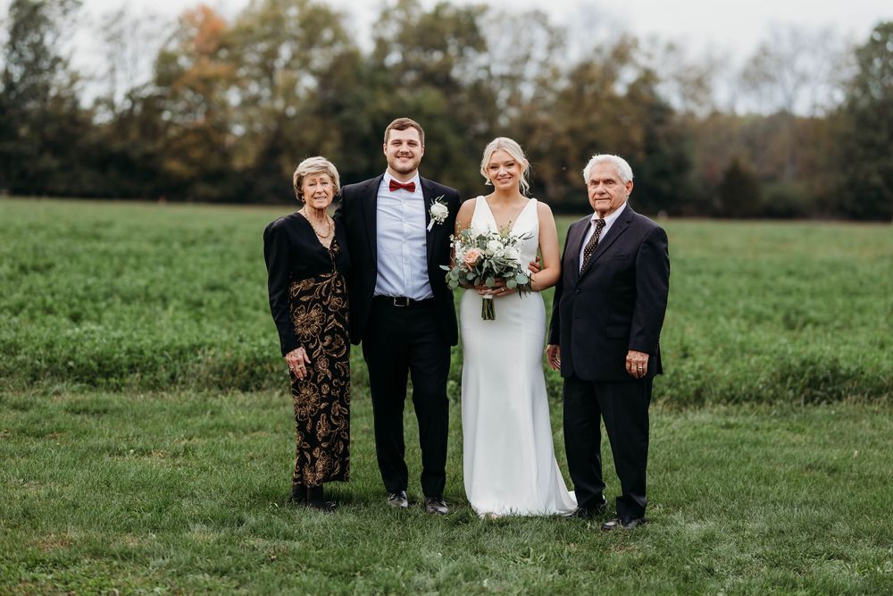 Groom in black tuxedo and bride in white gown posing with parents in green field at wedding.