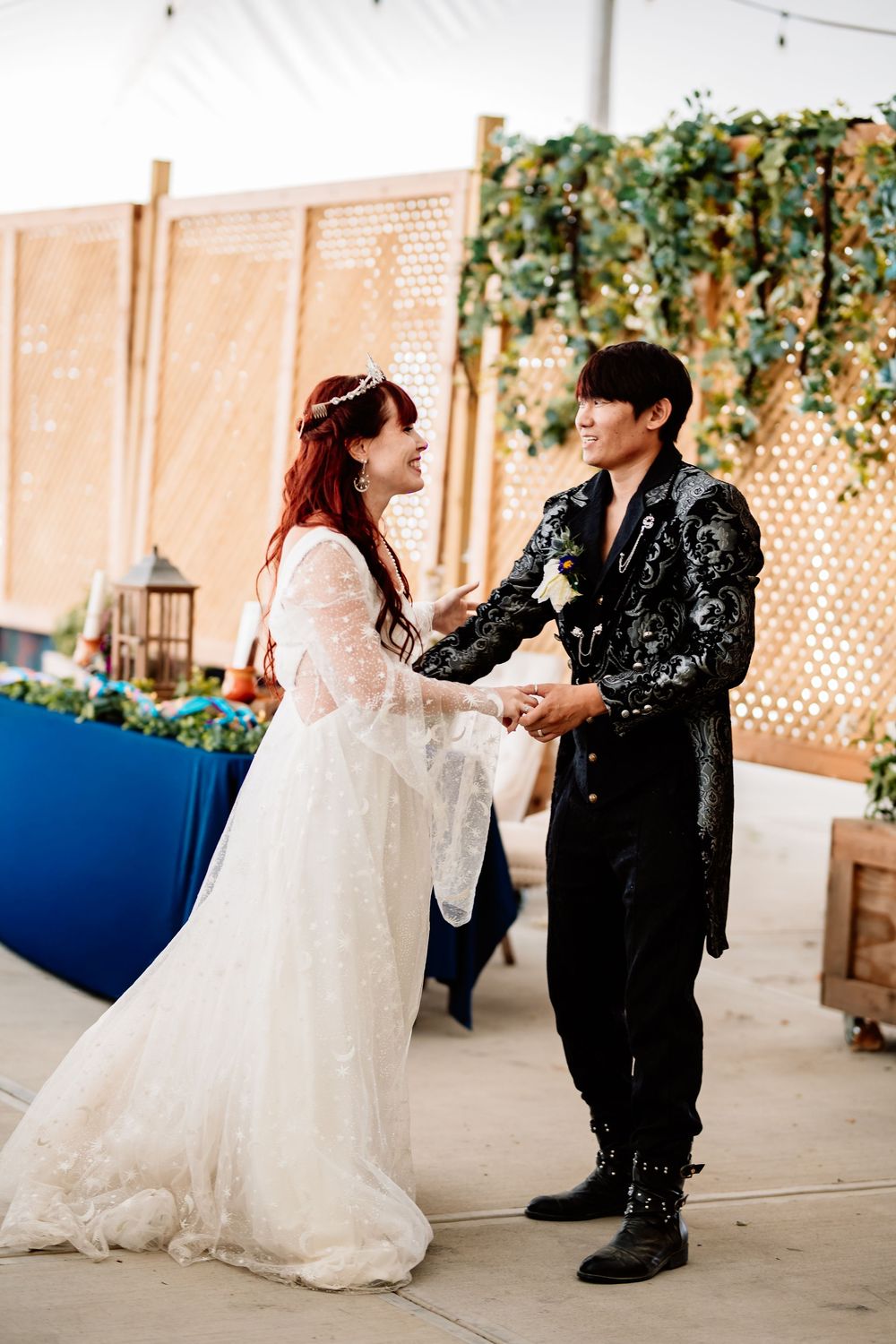 Bride in white lace gown and groom in black embroidered jacket hold hands smiling at wedding reception.