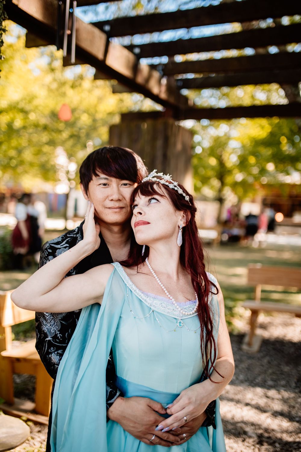 Couple in Renaissance-inspired outfits embrace under wooden pergola at outdoor medieval-themed wedding venue.
