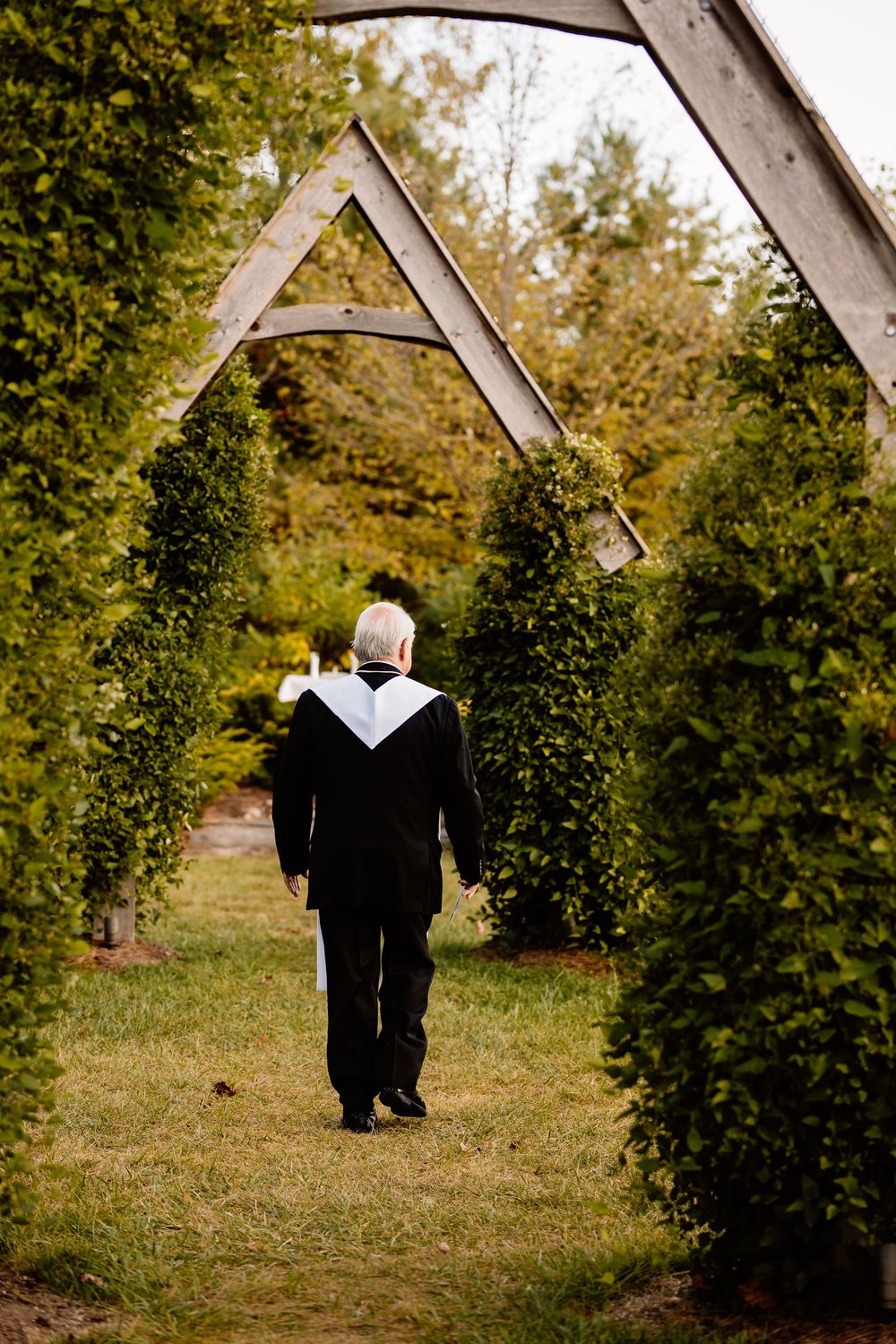 Officiant walks through a lush green hedge archway at an outdoor wedding ceremony.
