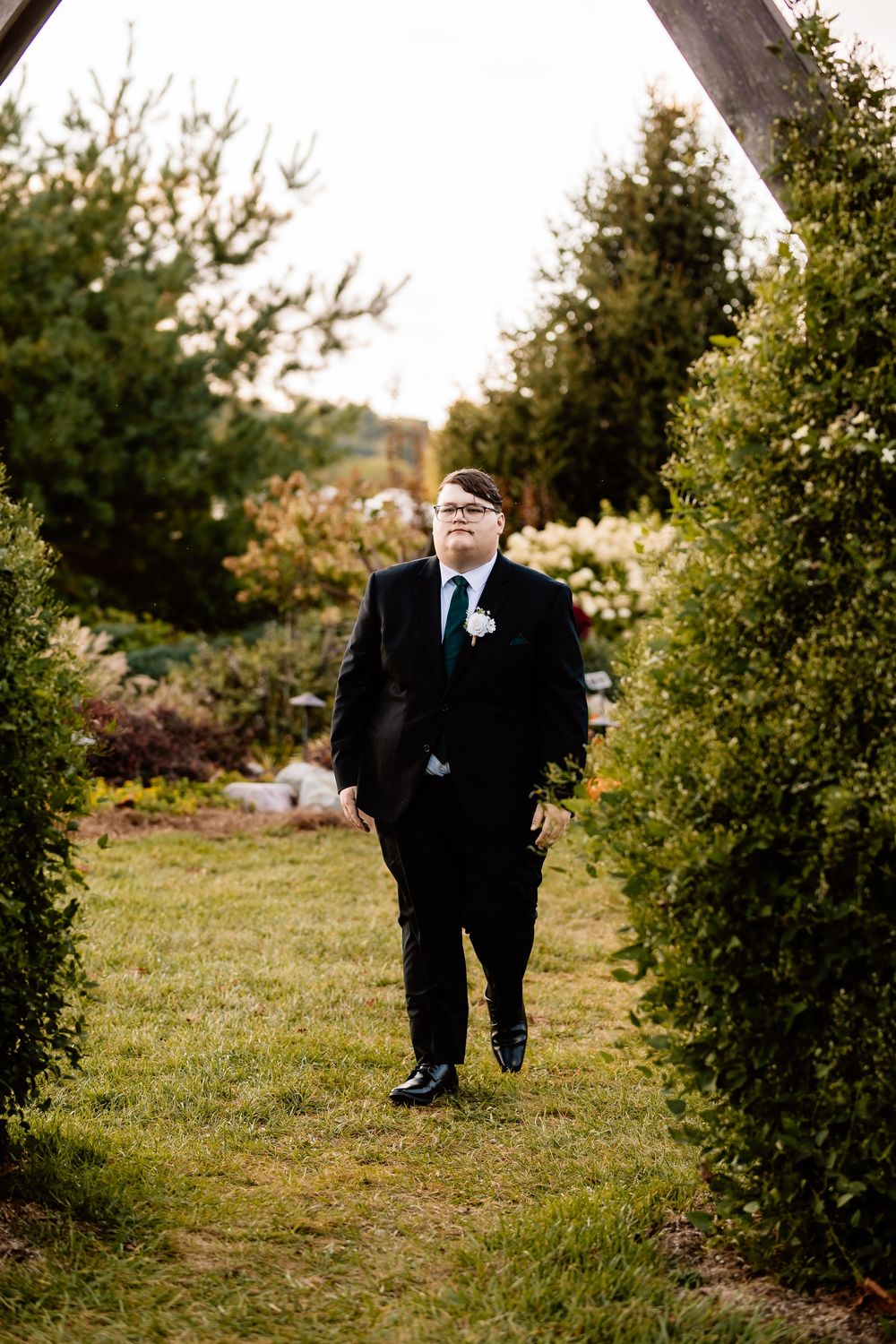 Groom stands confidently in a black suit under a wooden arch surrounded by green hedges.