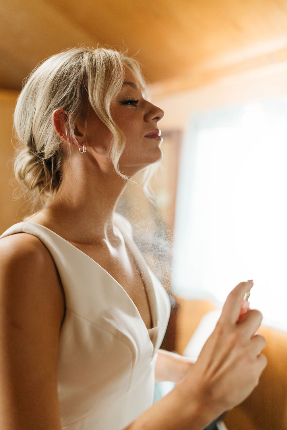 Blonde bride in white gown applying setting spray during wedding day preparations in warm light.