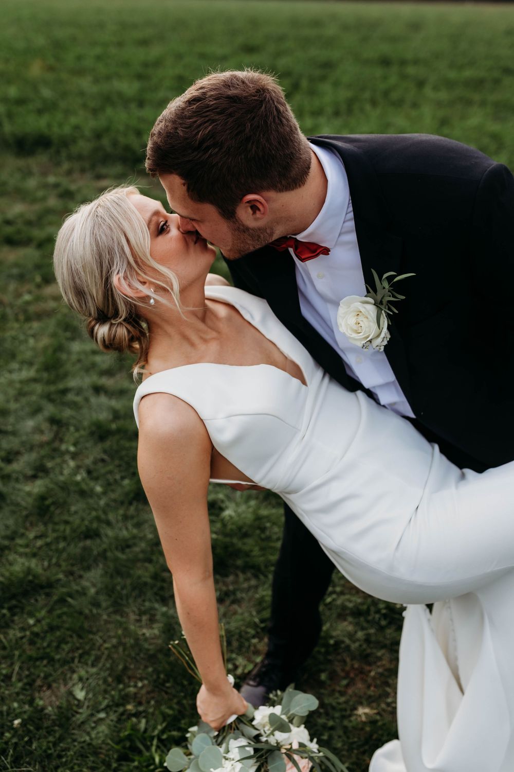 Groom dips bride for a romantic kiss outdoors on lush green grass at their wedding.