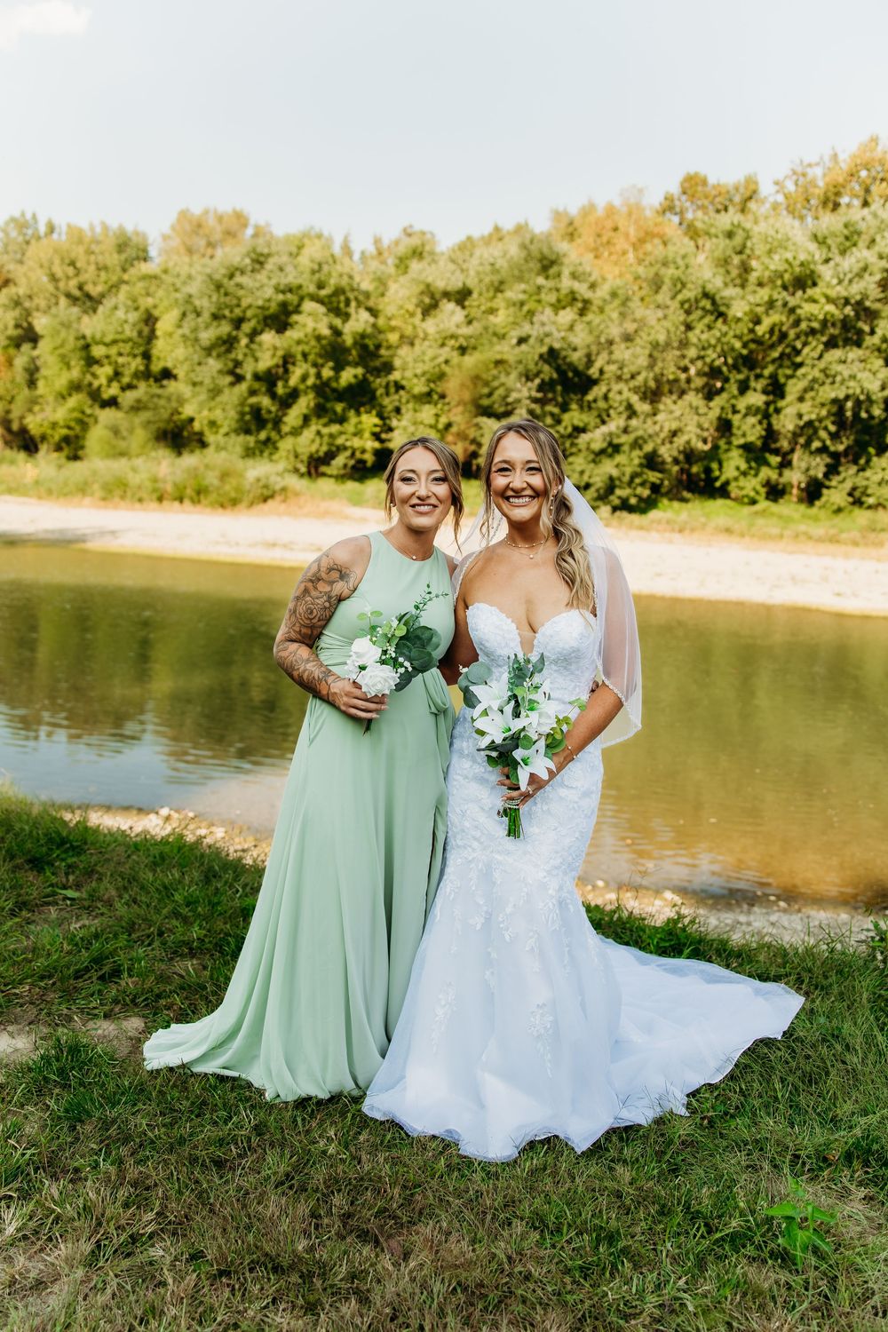 Bride in lace mermaid gown and bridesmaid in sage green dress pose by a scenic river.