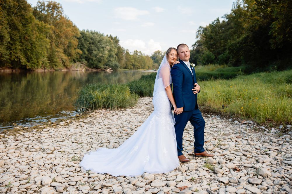 Bride and groom stand back-to-back on rocky riverbank surrounded by lush trees in golden light.