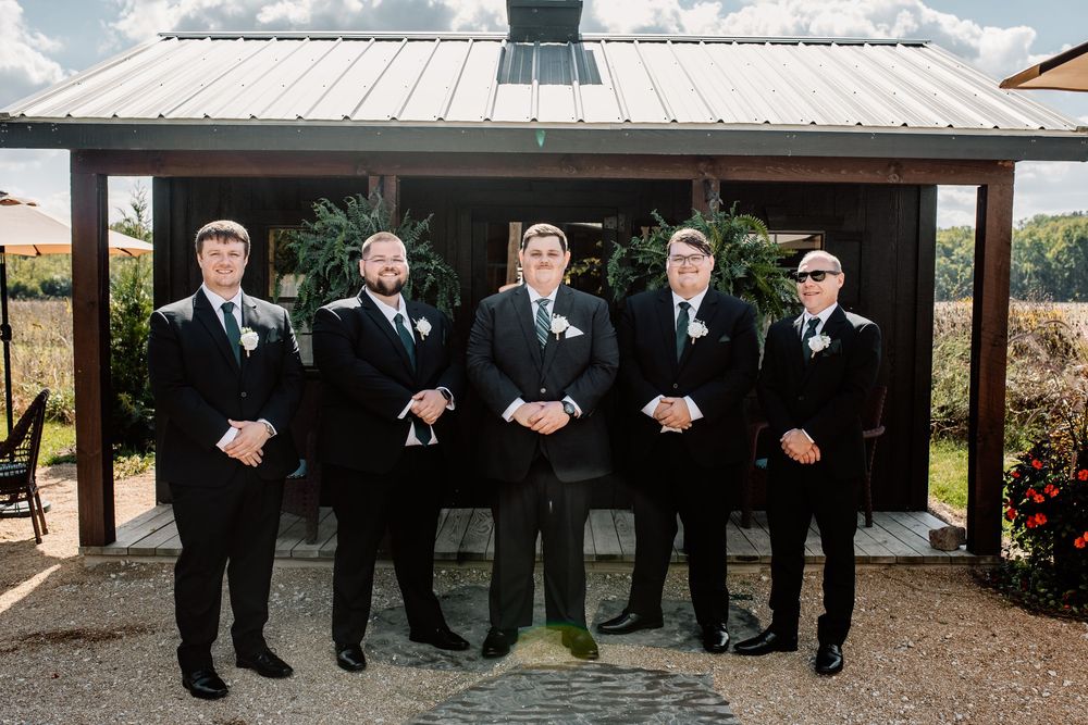 Five groomsmen in black suits with green ties pose together outside a rustic wedding venue.