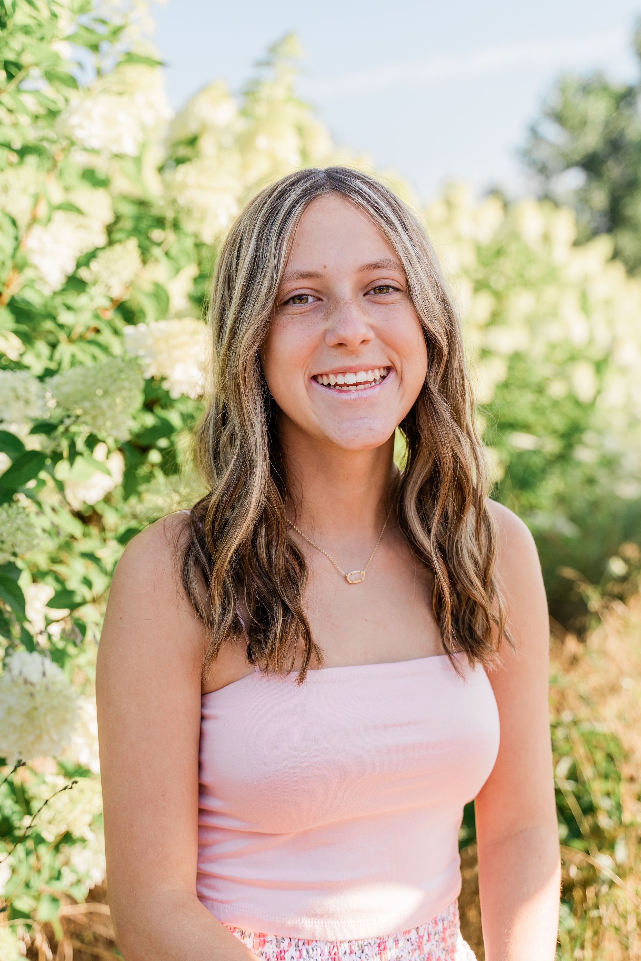 A person wearing a pink tank top and floral skirt smiles outdoors in front of green foliage on a sunny day.