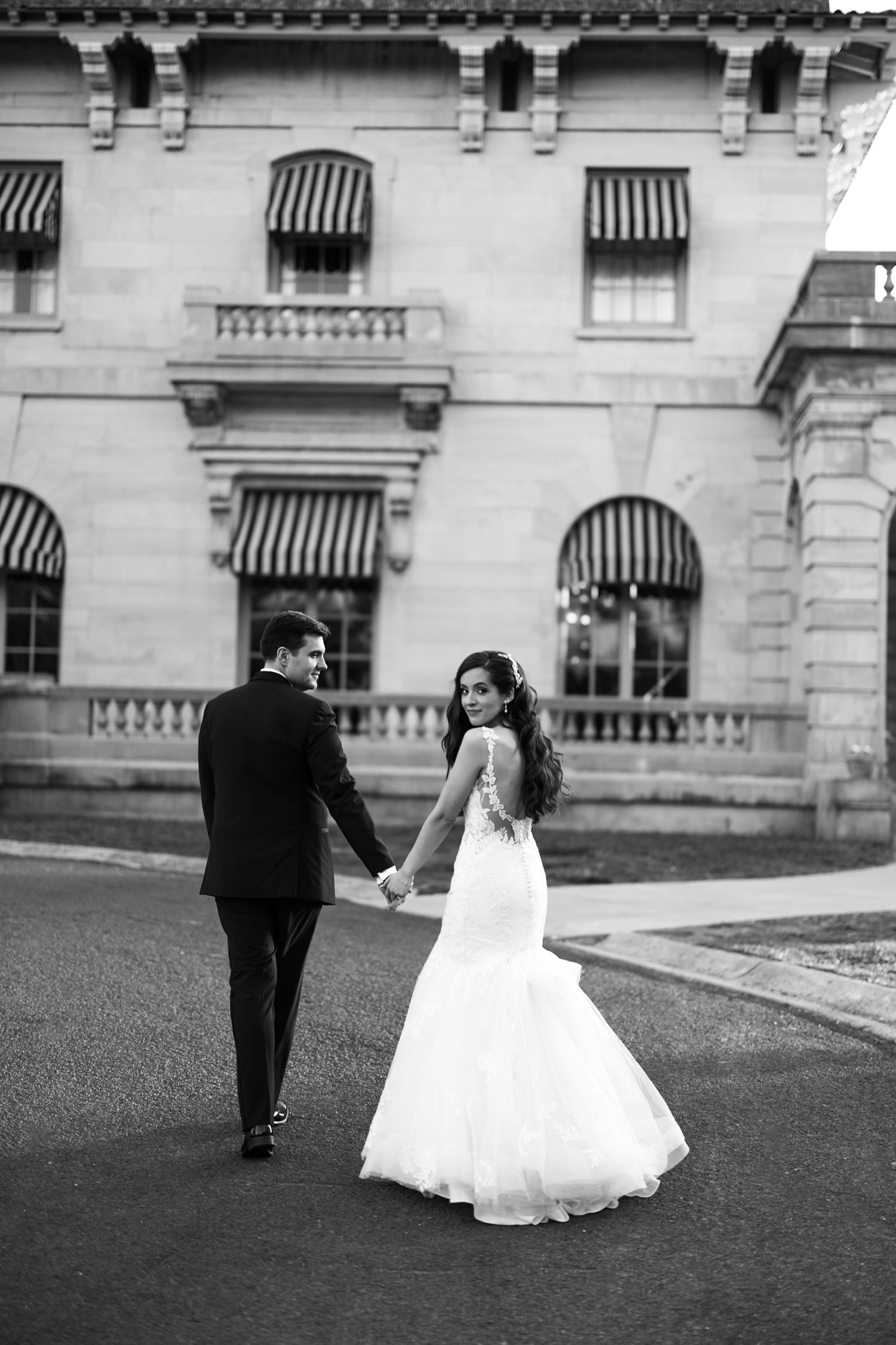 A couple in wedding attire walks together in front of a grand private mansion in loose editorial style and black and white dramatic editing.