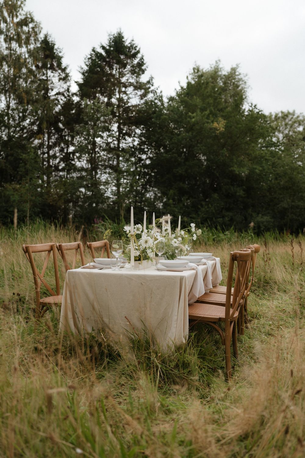 A rustic outdoor dining setup with wooden chairs and white tablecloths surrounded by tall grass and trees.