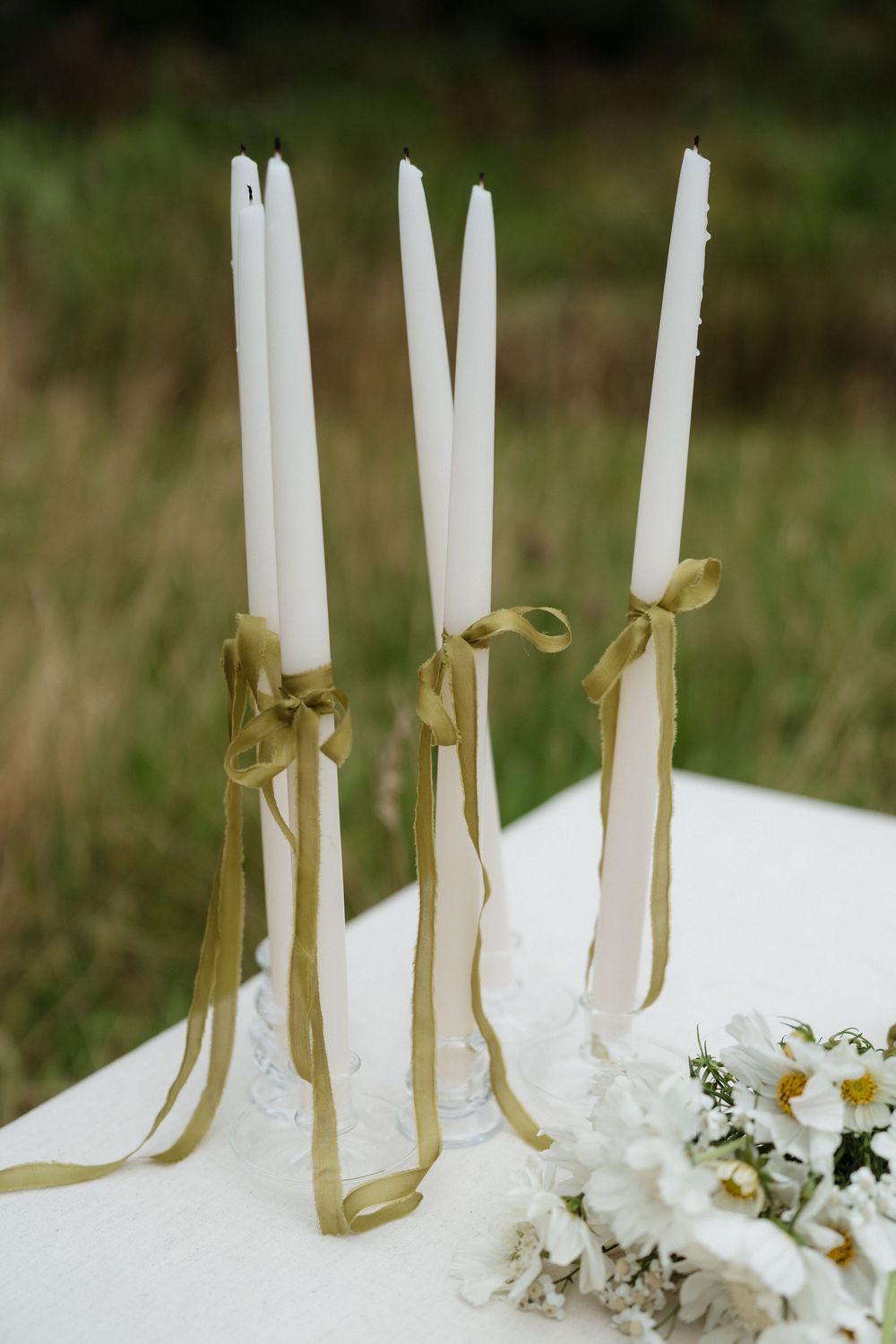 White taper candles with gold ribbons and delicate white flowers arranged on a blurred natural background.