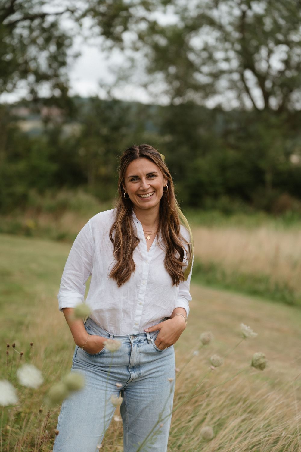 Person in white blouse and light jeans poses in a rustic field with tall grass and trees in the background.