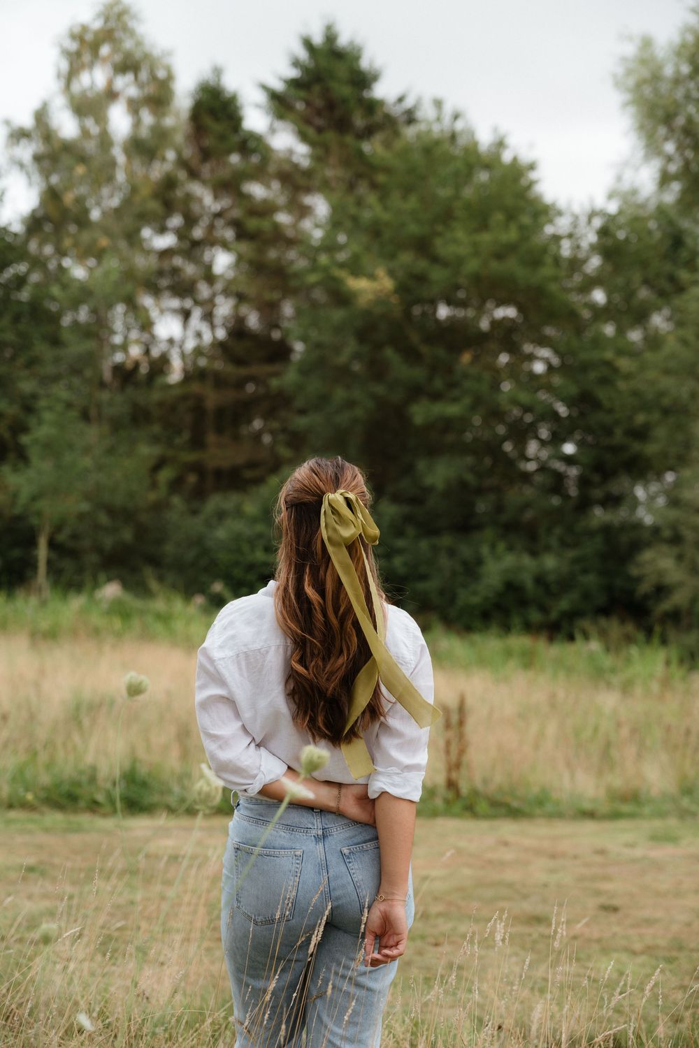 A person in denim overalls stands in a grassy field surrounded by trees and natural scenery.
