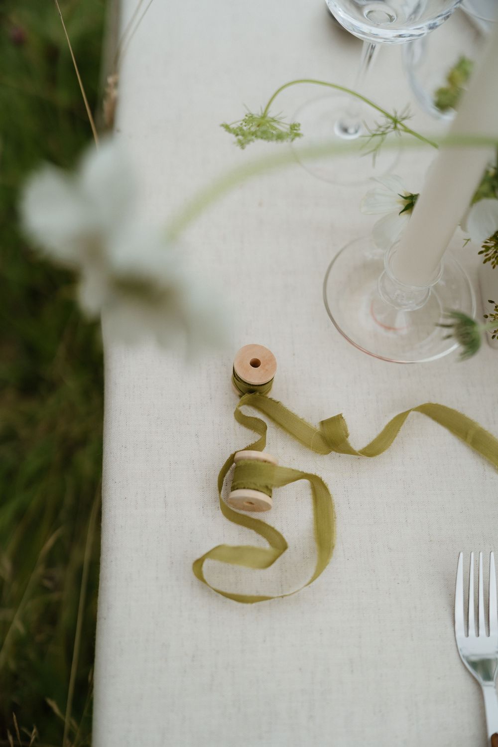 Elegant table setting detail with curled olive green ribbon and wine glass on white linen tablecloth.