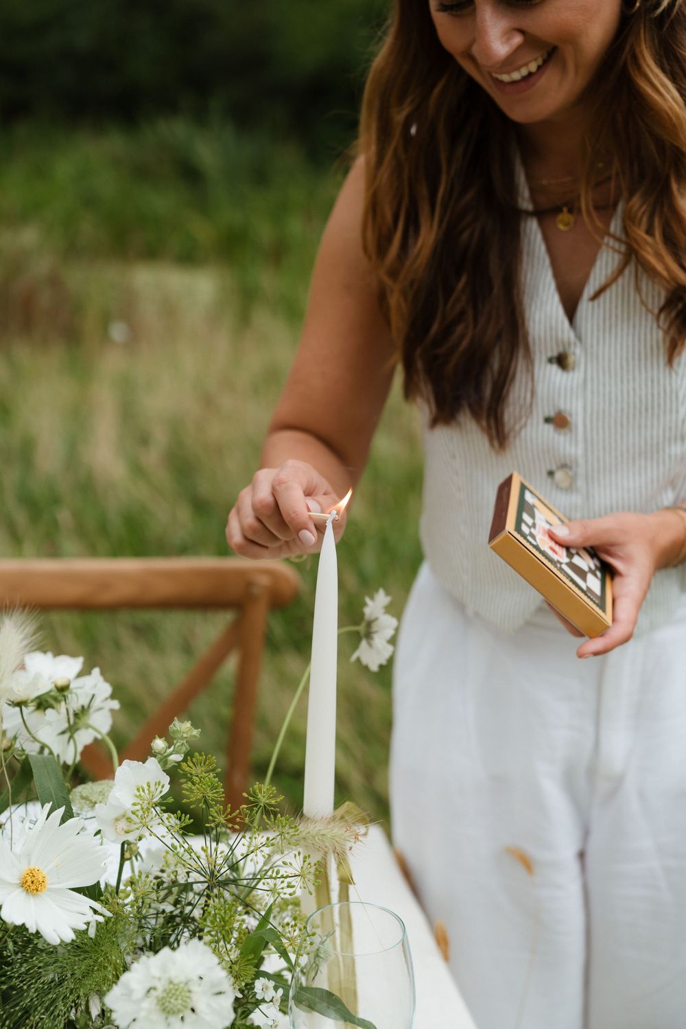Person in white dress lighting a candle among fresh white flowers and greenery in an outdoor setting.