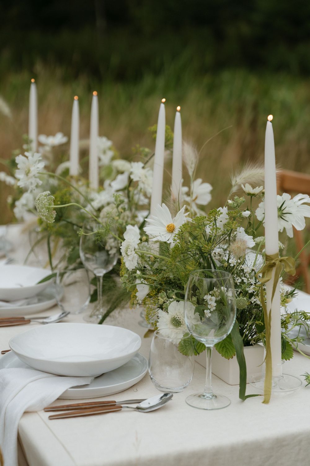 Elegant white tablescape with tall taper candles and delicate greenery arrangements on a crisp white tablecloth.