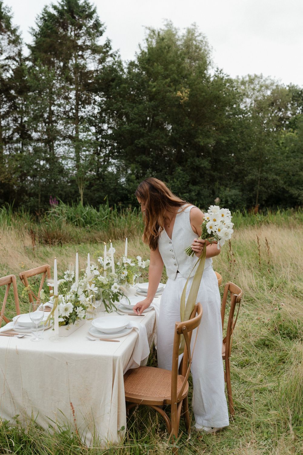 Rustic outdoor dining setup with white tablecloth and wooden chairs in a natural meadow setting.