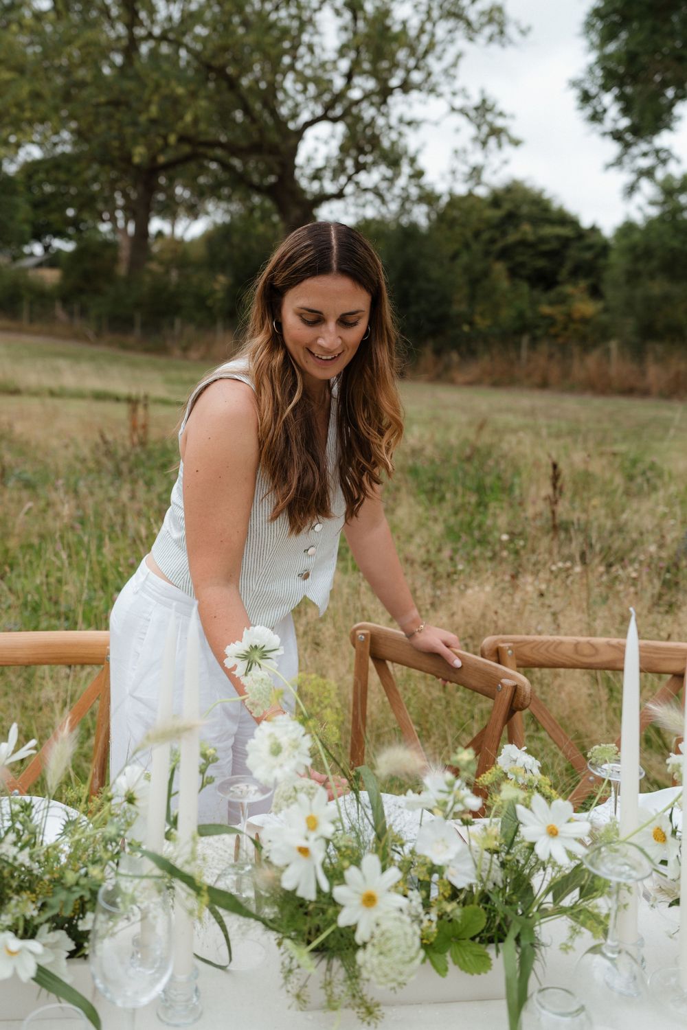 A person in a white dress arranges delicate white flowers along a rustic wooden fence in a natural outdoor setting.