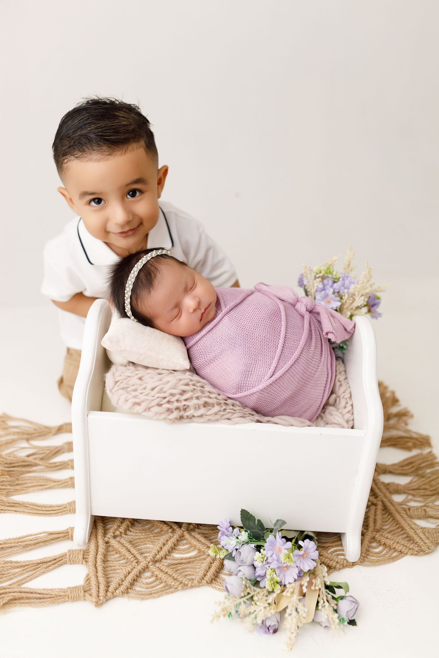 Sweet newborn photography scene with baby wrapped in pink on white wooden bed with purple flowers and sibling nearby.