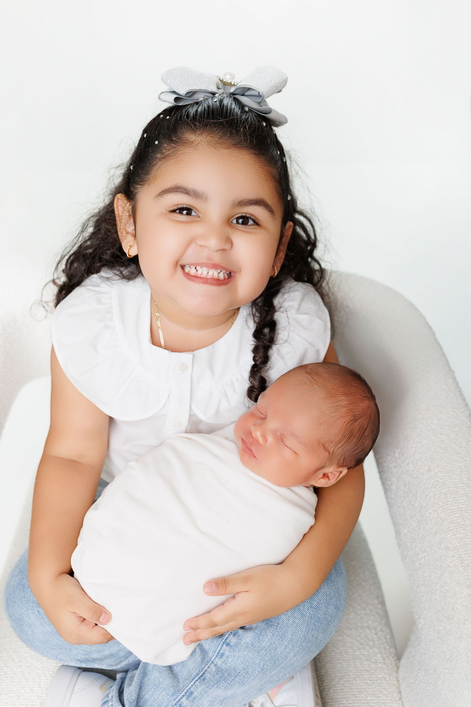 Young girl in white dress sits with newborn baby in series of tender sibling photos against white background.