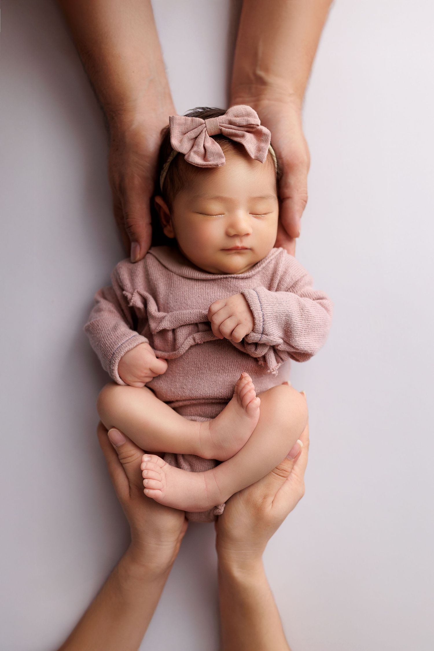 Peaceful sleeping baby wearing pink outfit and bow headband cradled by gentle hands on white background.