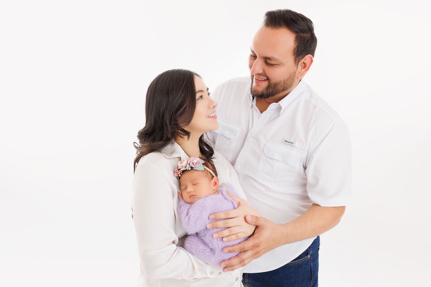 Expecting couple in white outfits sharing tender moments during maternity photoshoot against white background.