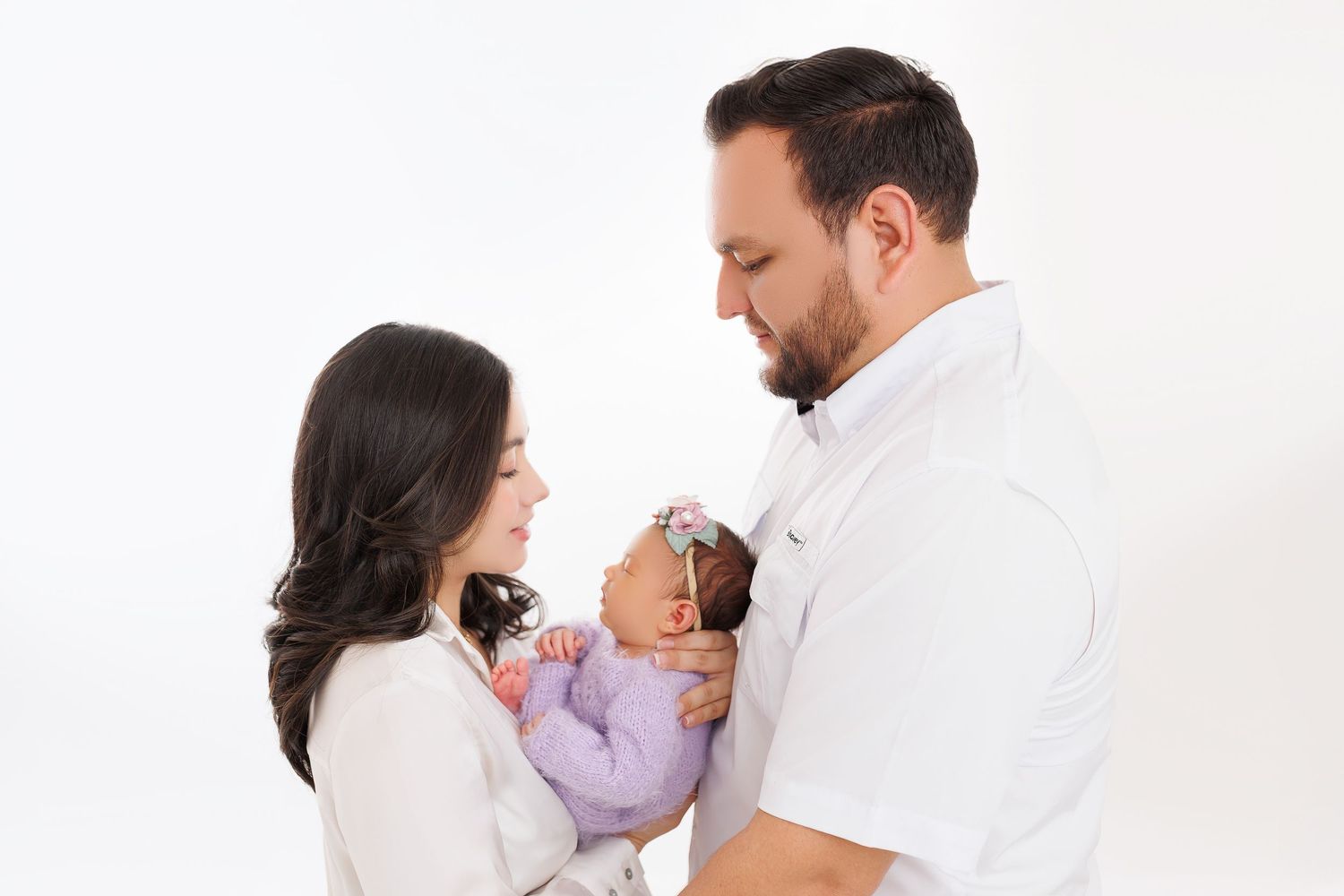 Parents in white outfits share a tender moment with their newborn baby wearing purple against a white backdrop.