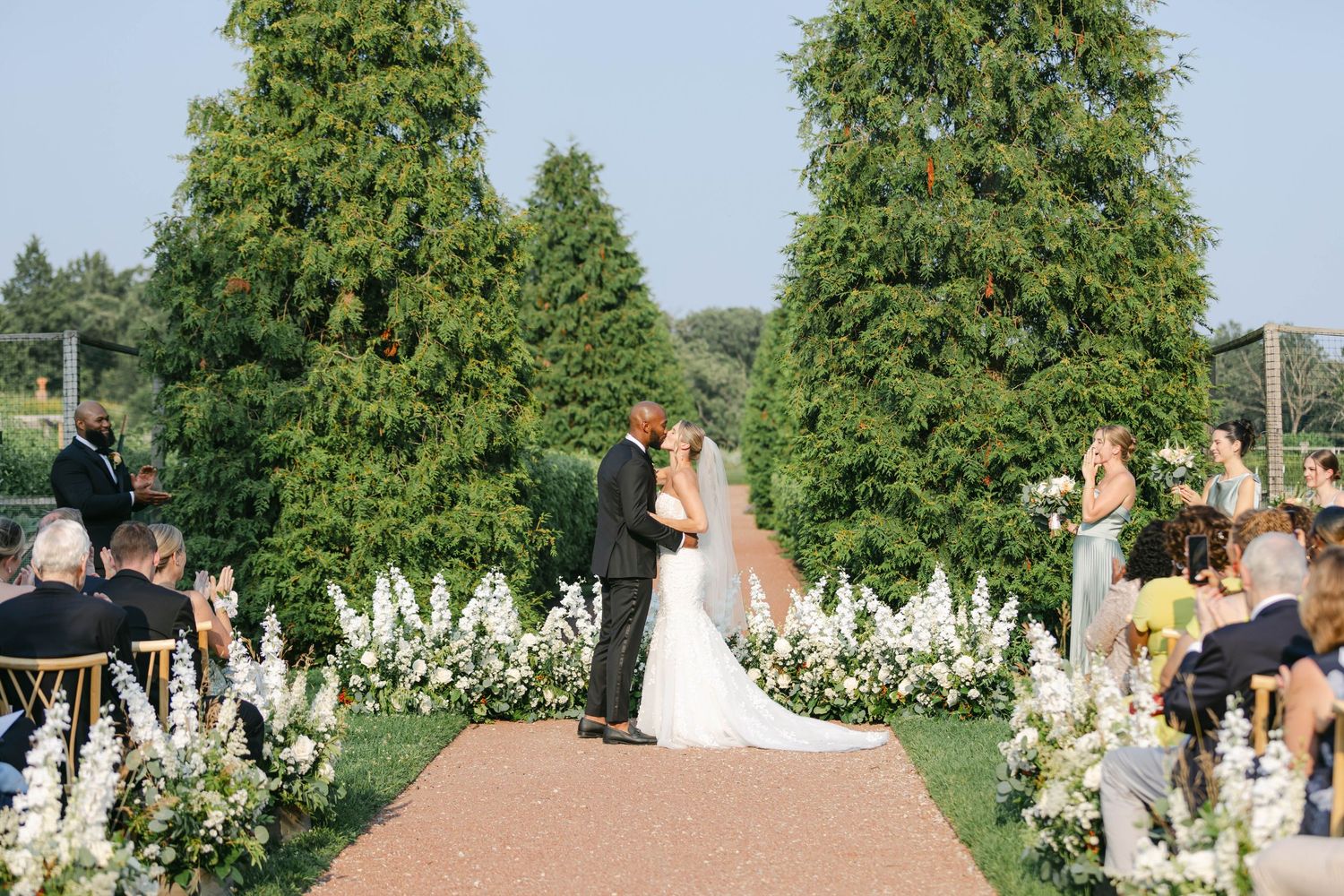 Couple shares intimate moment in lush garden setting surrounded by white flowers and greenery. - Elawa Farm