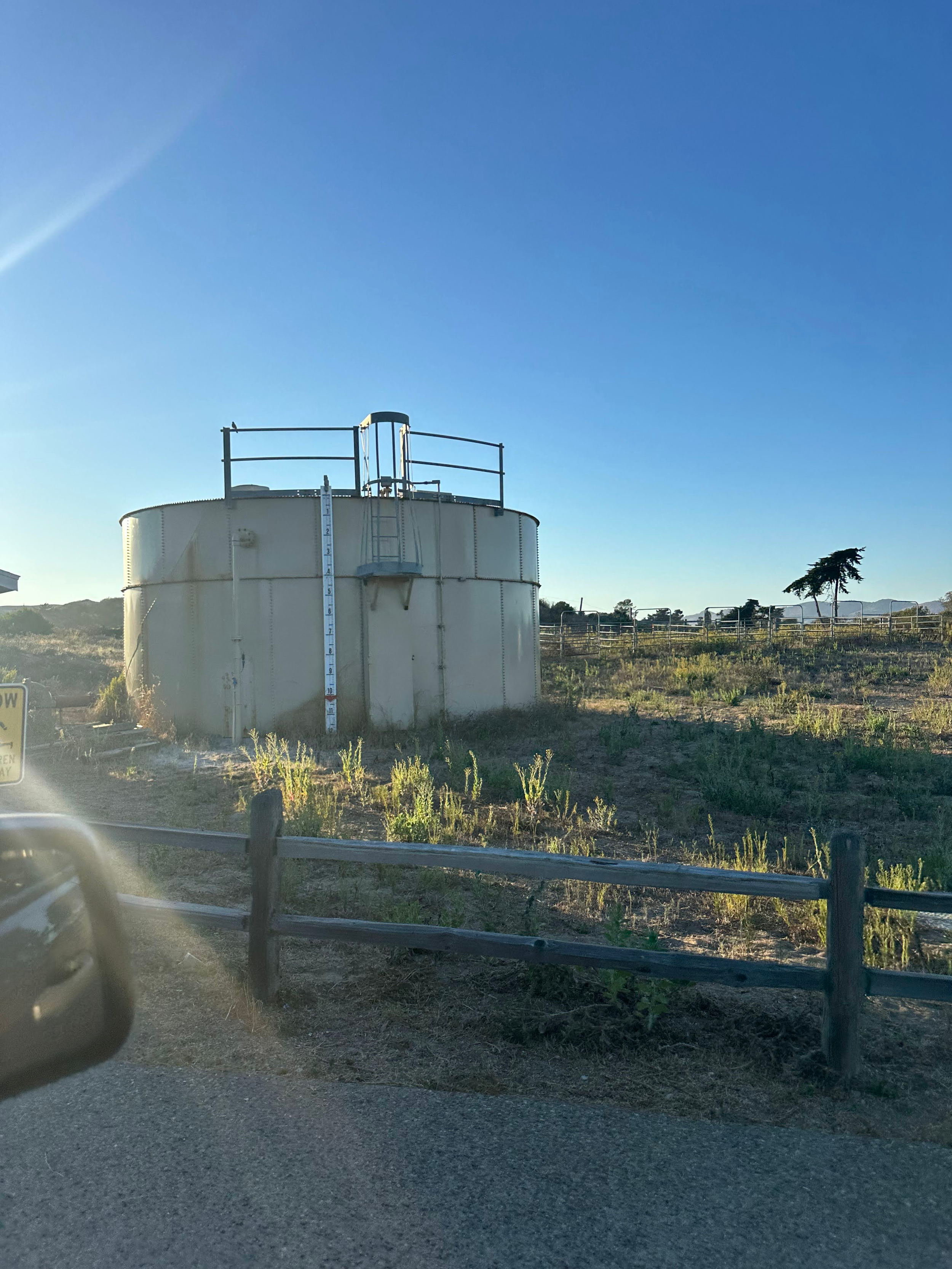 A large white storage tank with metal railing sits in an arid landscape beside a dirt road.