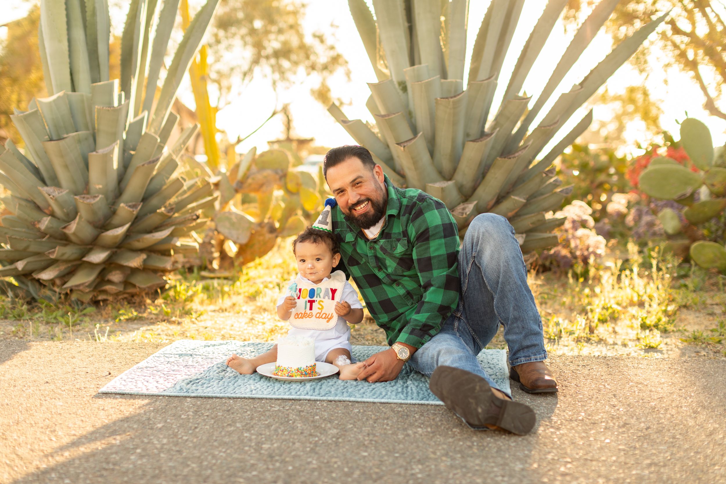 Parent and child sharing special moment with cake during outdoor photoshoot near agave plants.