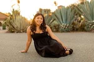 A person in a black dress poses on the ground with agave plants and sunset lighting in the background.