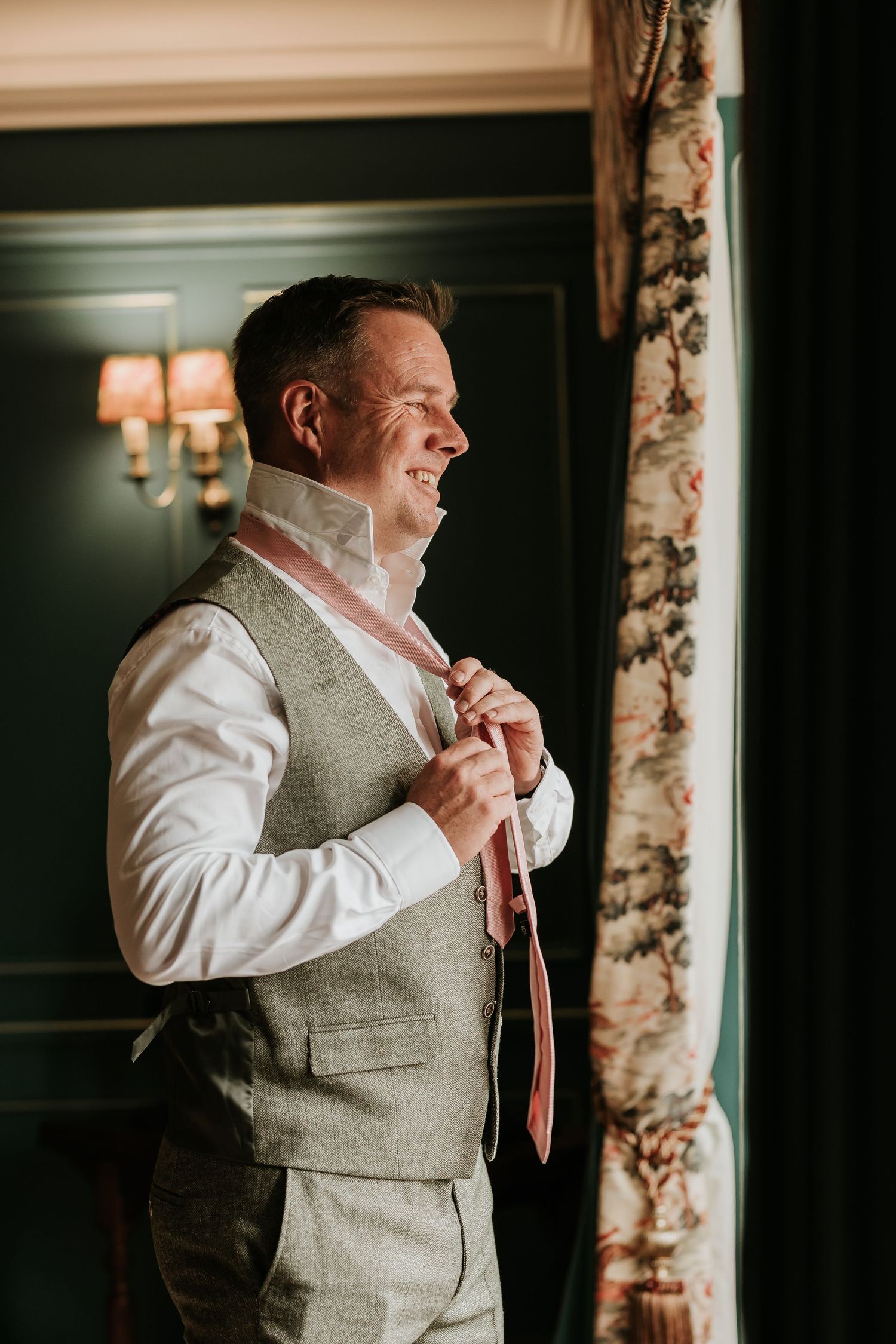 A person in a white shirt and gray vest adjusts their tie while smiling in a warmly lit room with floral decor.