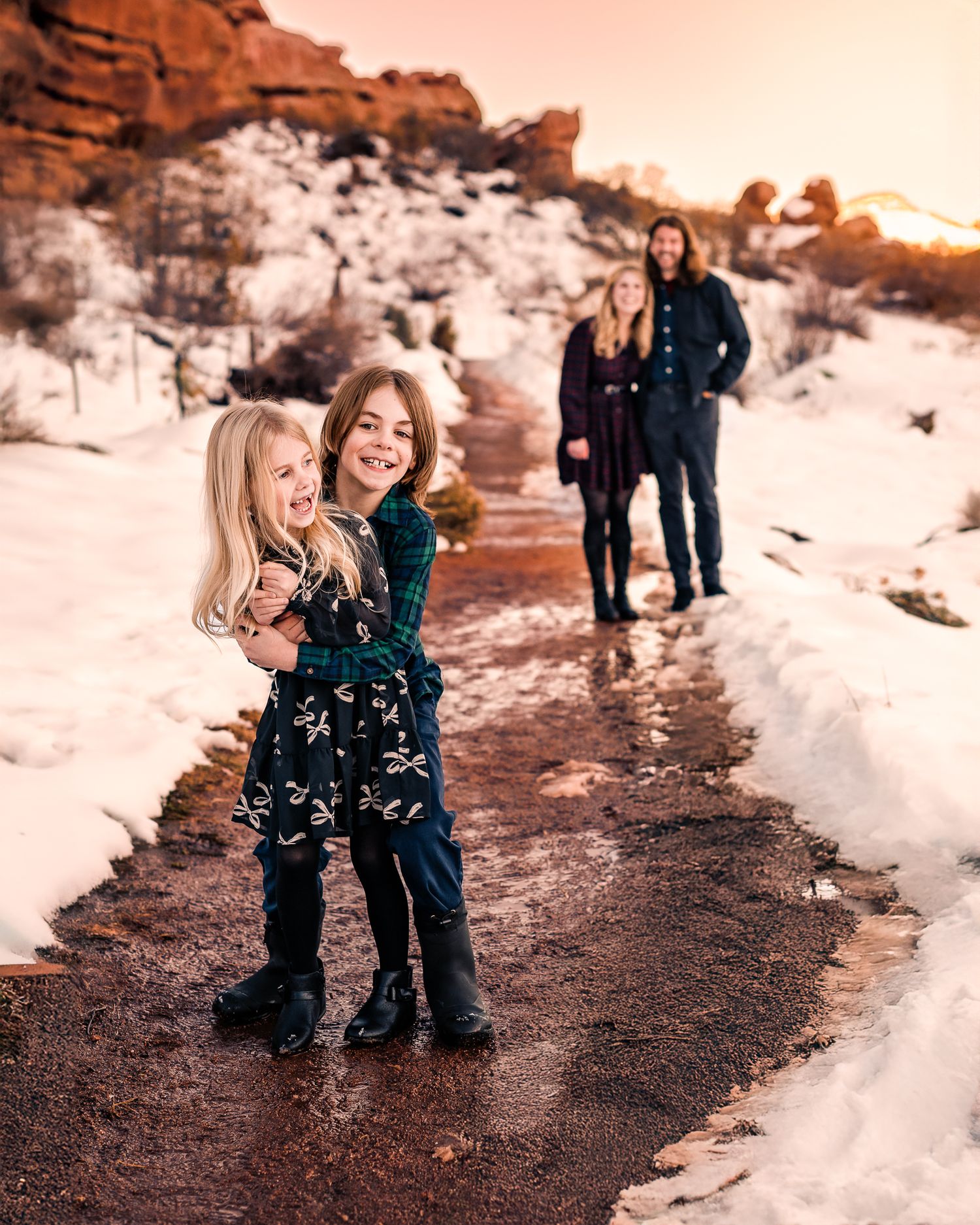 A brother and sister playfully hugging while a mom and dad look on amongst the red rocks in Morrison, CO