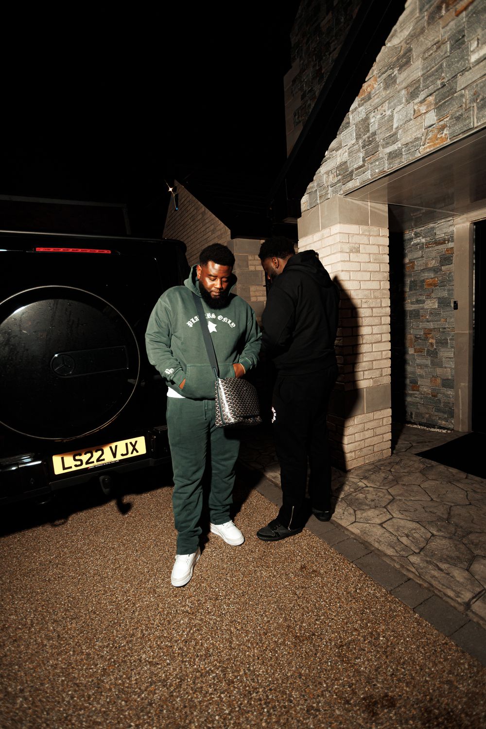 A person in a green uniform walks through a dimly lit parking area at night casting shadows on brick walls.