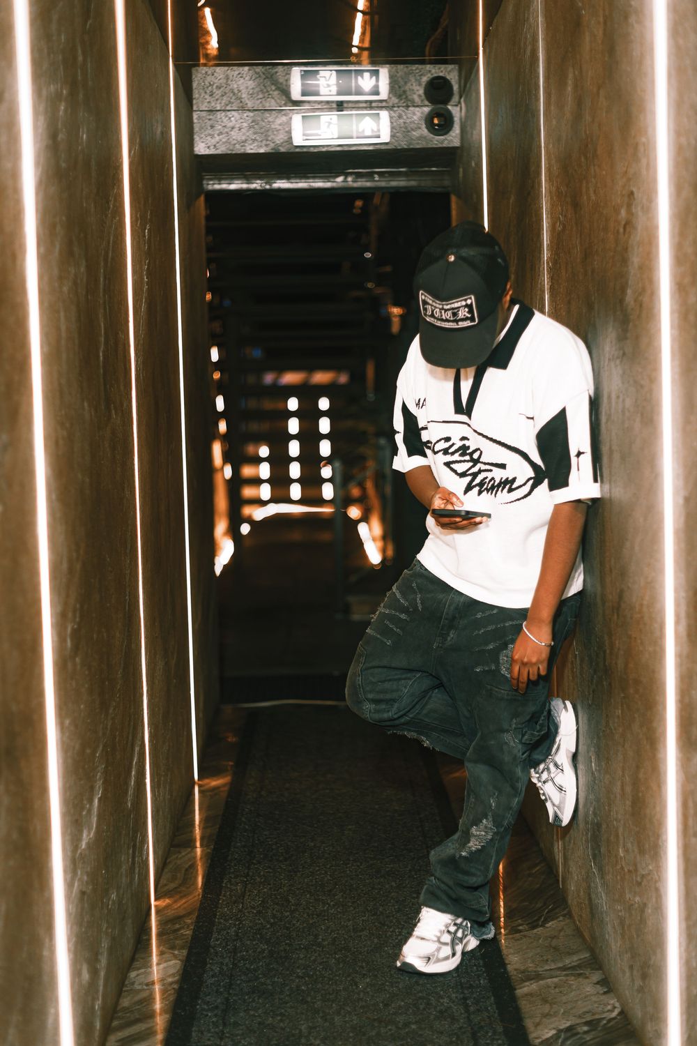 Person wearing a white t-shirt and black cap poses in a dimly lit hallway with metal walls.