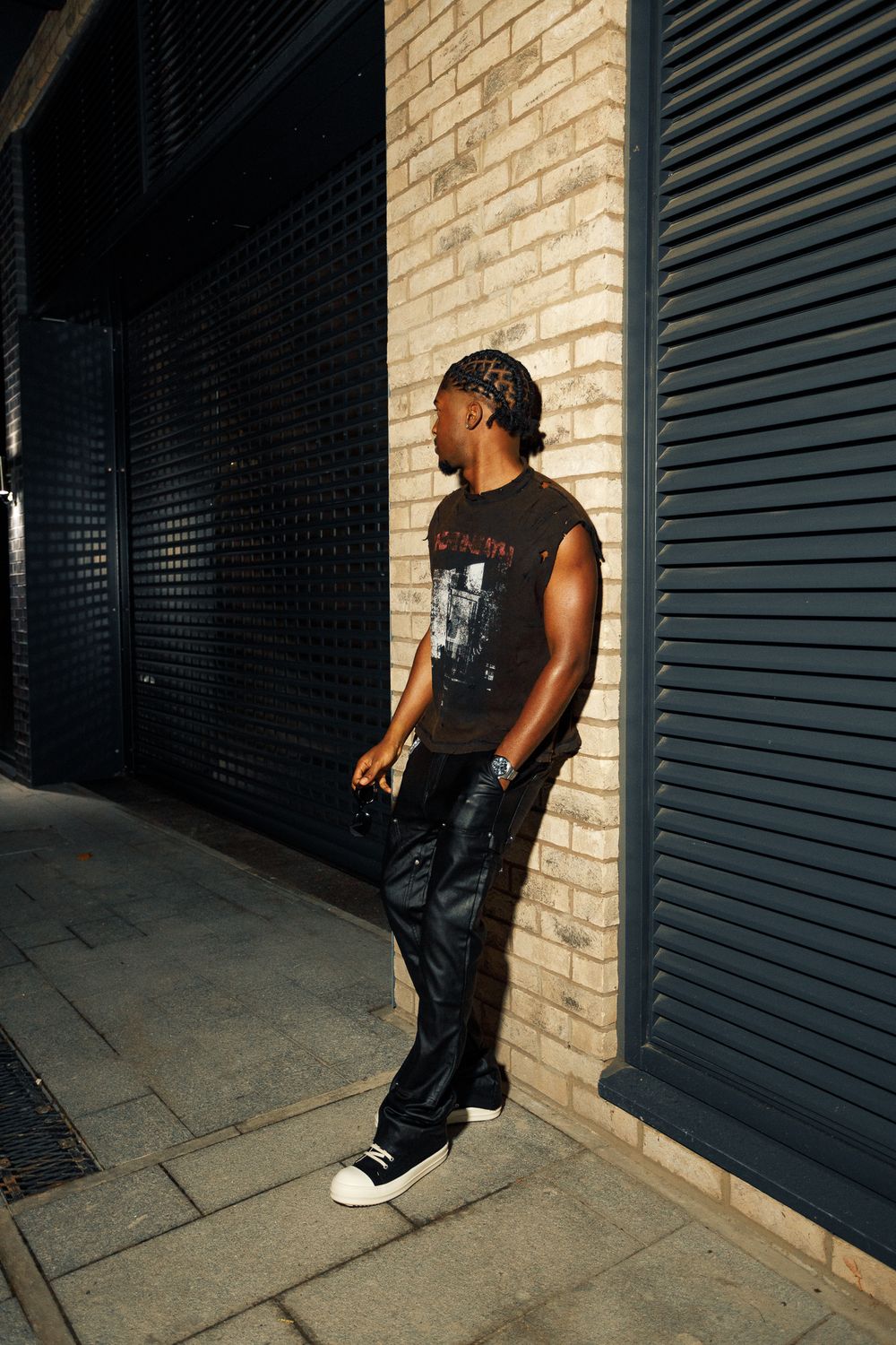 Person in black sleeveless shirt and dark jeans leans against brick wall in urban setting with dramatic lighting.