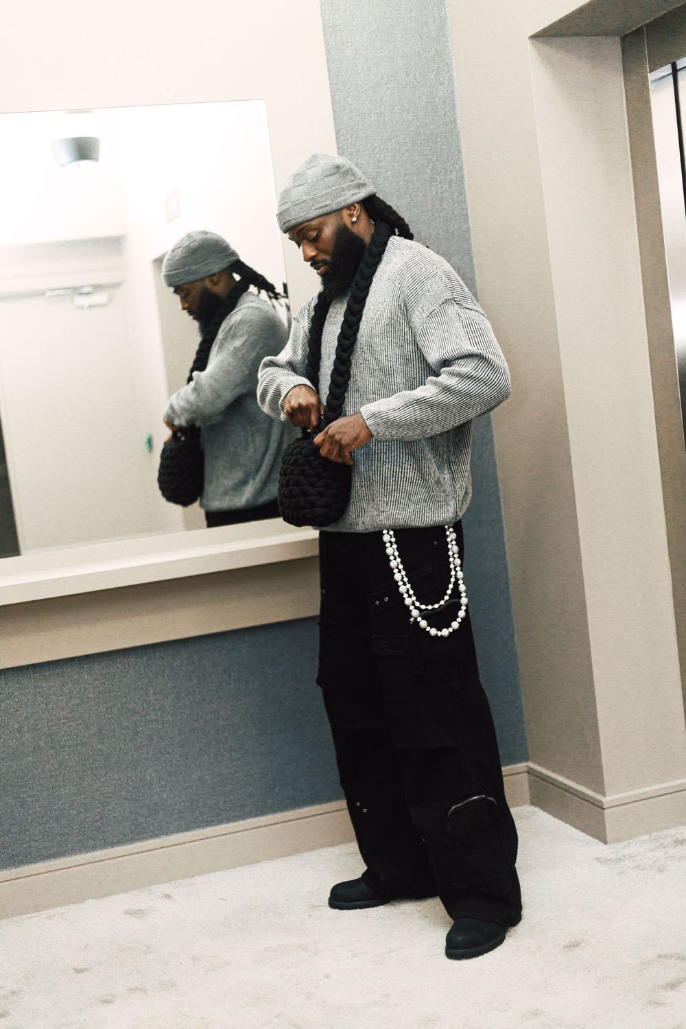 Person in grey hoodie and beanie with baggy black pants and chain accessory poses in front of mirror.