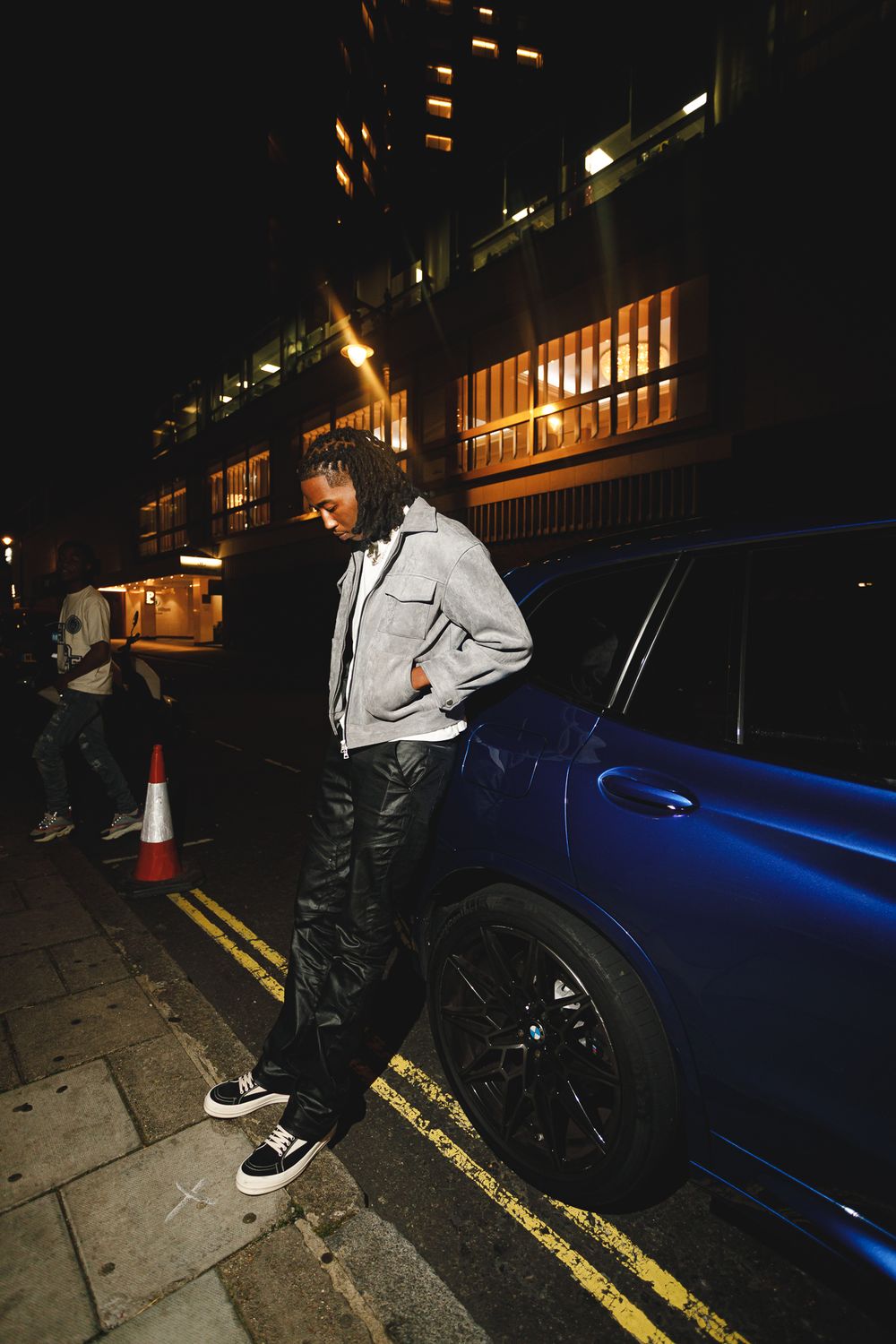 Person in a light colored jacket and dark pants standing next to a blue car on a nighttime city street.